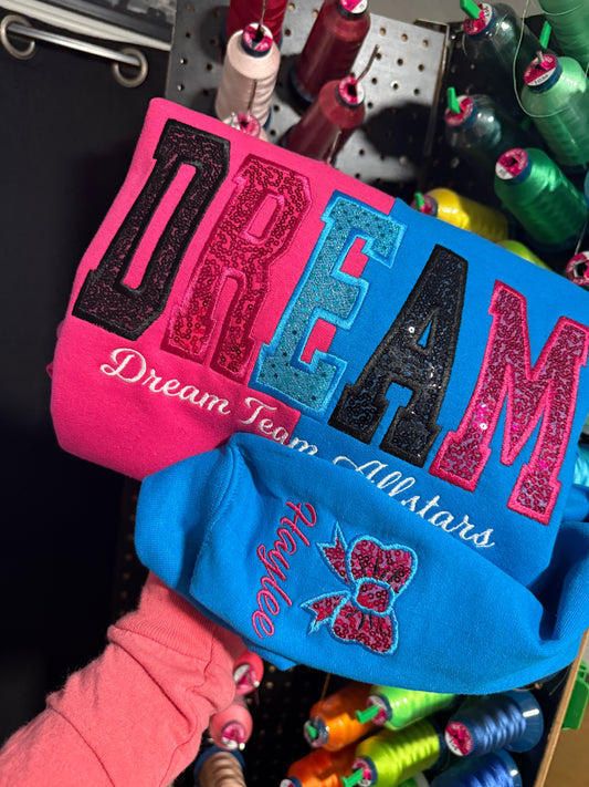 Two embroidered sweatshirts, one pink and one blue, with text and graphics on a background of spools of thread.